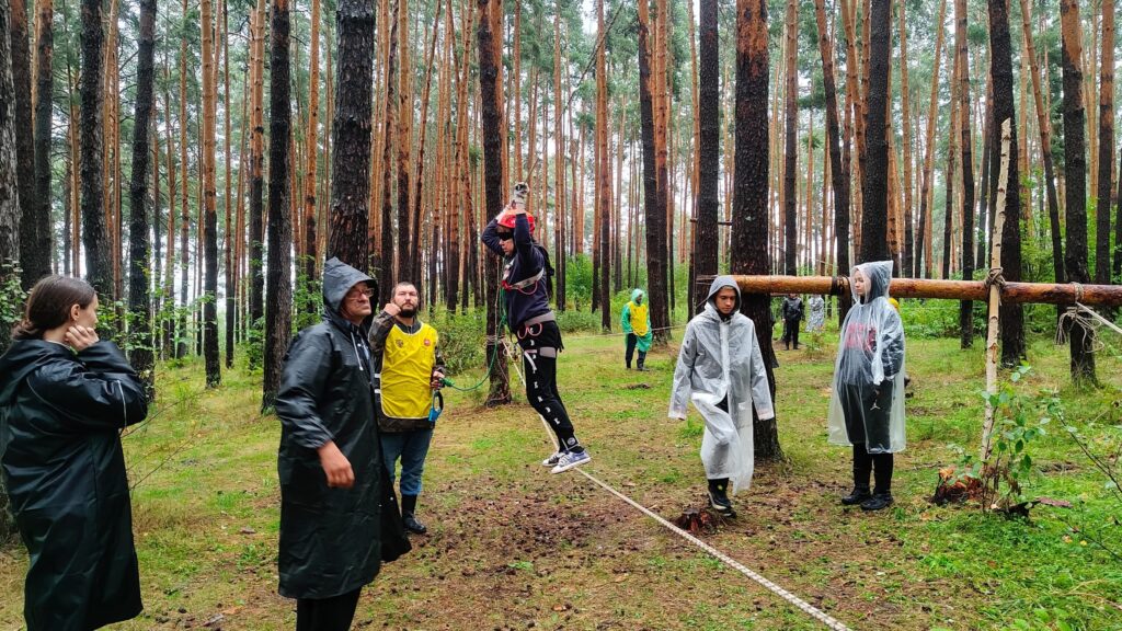 На фото участники туристических сборов проходят этап полосы препятствий в лесу — человек в страховке и каске передвигается по верёвке, а остальные наблюдают и страхуют. Погода дождливая, многие в дождевиках.