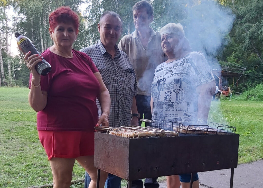 На фото — группа людей на природе у мангала с решётками, на которых готовится еда. На переднем плане женщина в красной футболке и шортах держит бутылку маринада или соуса, рядом с ней ещё трое участников встречи. Вокруг — зелёная поляна и деревья.