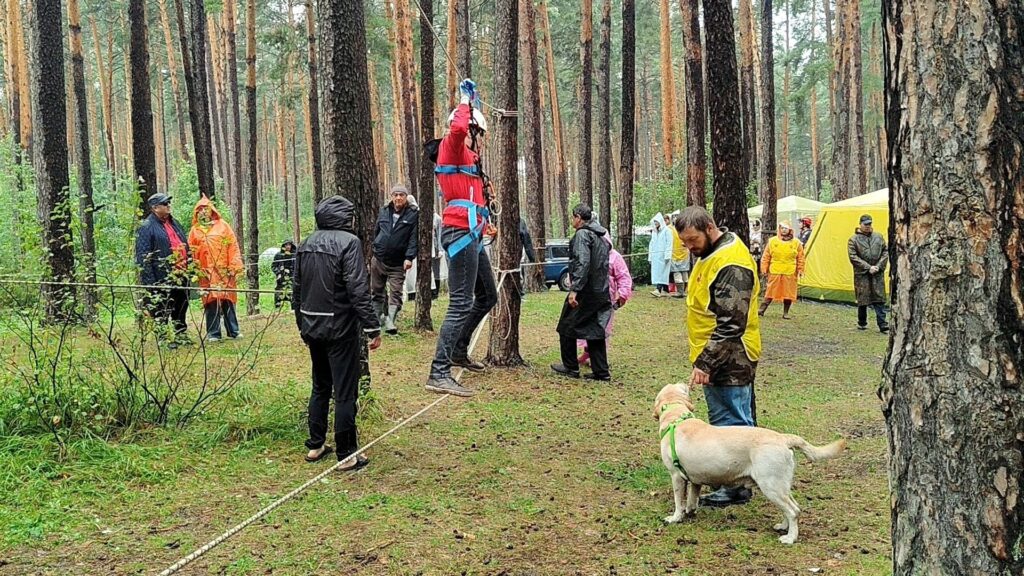 Спортивный этап с подъёмом по верёвке, рядом — участник с собакой-проводником, наблюдающие за происходящим