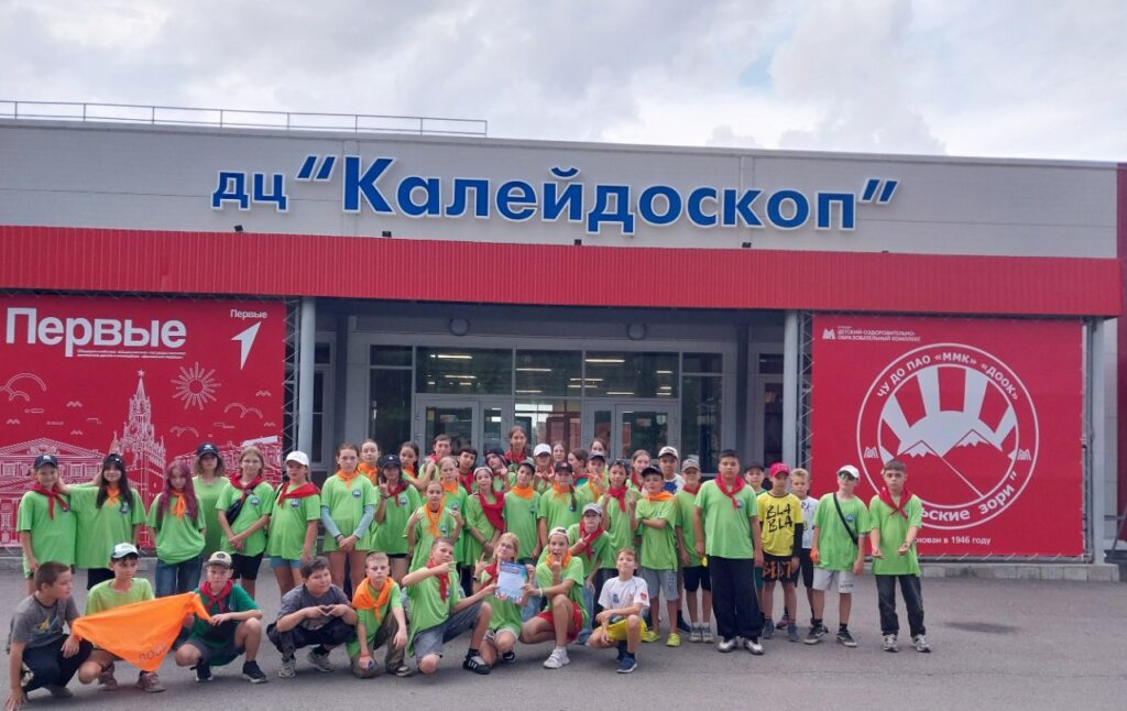 Участники детской команды у ДЦ «Калейдоскоп»
Группа ребят в ярких зелёных футболках и галстуках позирует перед входом в детский центр.
