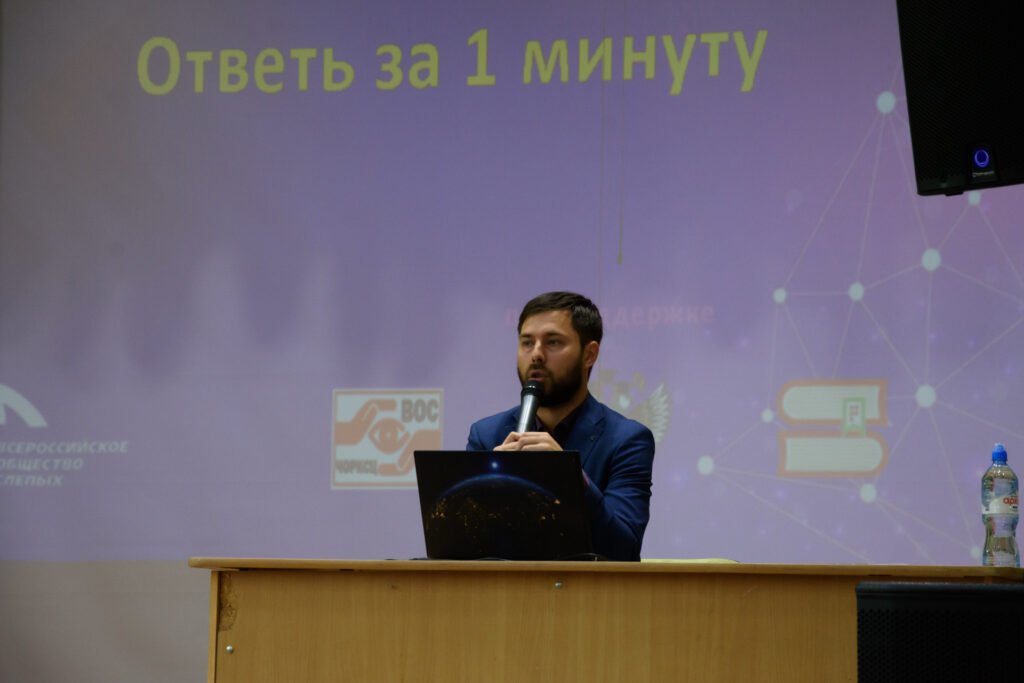 Ведущий турнира «Ответь за 1 минуту» задаёт вопросы командам. На фоне экрана с названием конкурса он держит микрофон, готовясь объявить следующий раунд.
