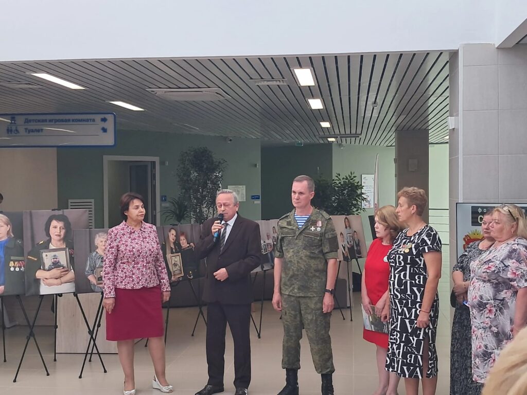 Момент открытия выставки. На переднем плане выступает мужчина в пиджаке с микрофоном, рядом — женщина в цветастой блузке. На фоне — военнослужащий, женщины, посетители и портреты семей героев на стендах.