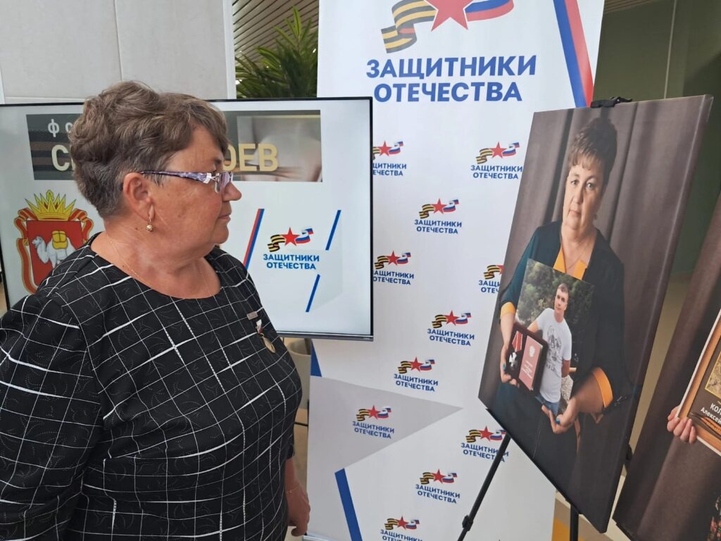 Пожилая женщина в очках смотрит на фотопортрет на стенде. На фото — женщина с серьёзным лицом держит фото молодого мужчины и награды. Позади баннер с надписью «Защитники Отечества».