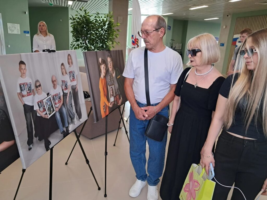 Три посетителя — мужчина и две женщины в чёрной одежде — рассматривают фотографии на выставке. На стендах — портреты родственников бойцов с наградами и детскими руками.