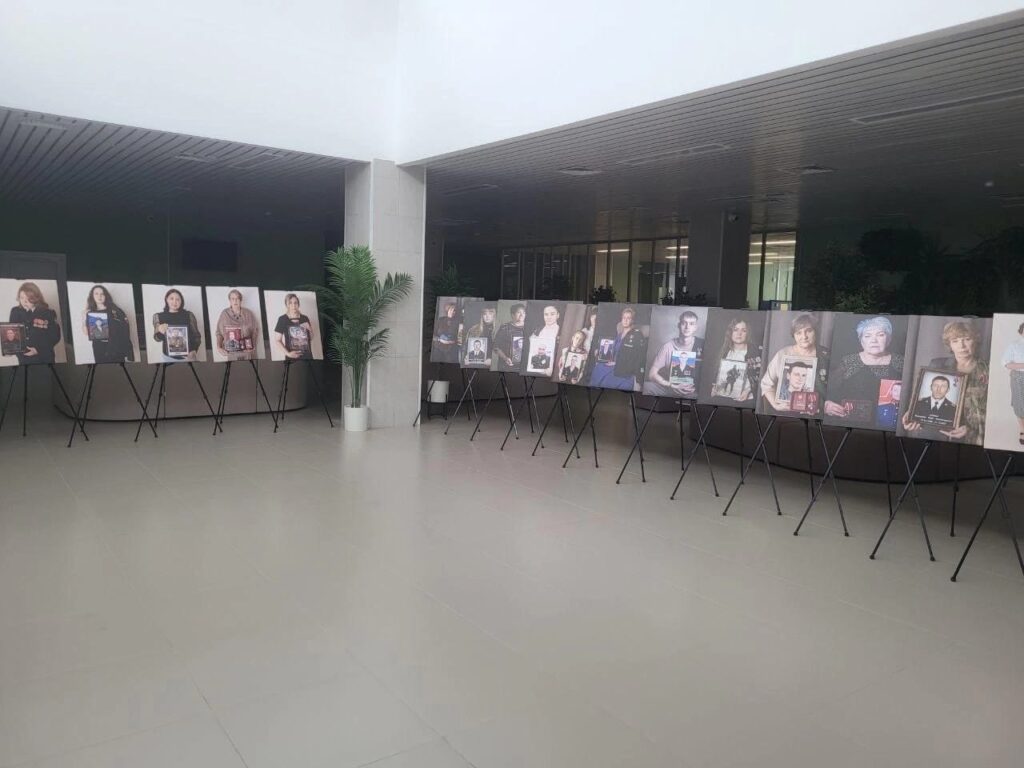 Панорамный вид зала. По стенам выстроены более двадцати фотопортретов участников проекта: женщины, мужчины и дети держат фотографии своих близких — героев СВО. Светлый зал, выставка оформлена в едином стиле.