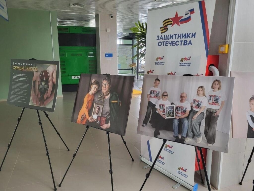 Три фотостенда на выставке. Слева — щит с названием «Семьи героев» и текстом о выставке. В центре — портрет женщины и мальчика с наградами и фотографией бойца. Справа — большая фотография семьи в футболках с портретом погибшего, позирующей на белом фоне.