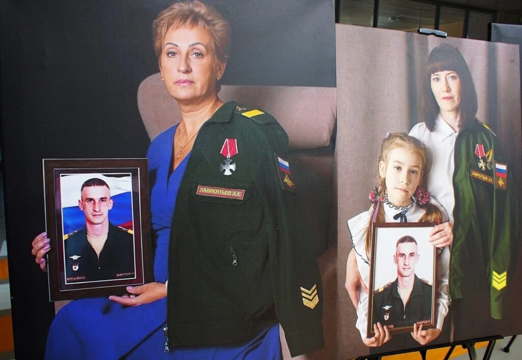 Два портрета на выставке. Слева — женщина в синем платье держит фото сына в военной форме и награды, рядом висит его китель. Справа — женщина и девочка с таким же кителем и фотографией того же бойца. Семейная память о погибшем.
