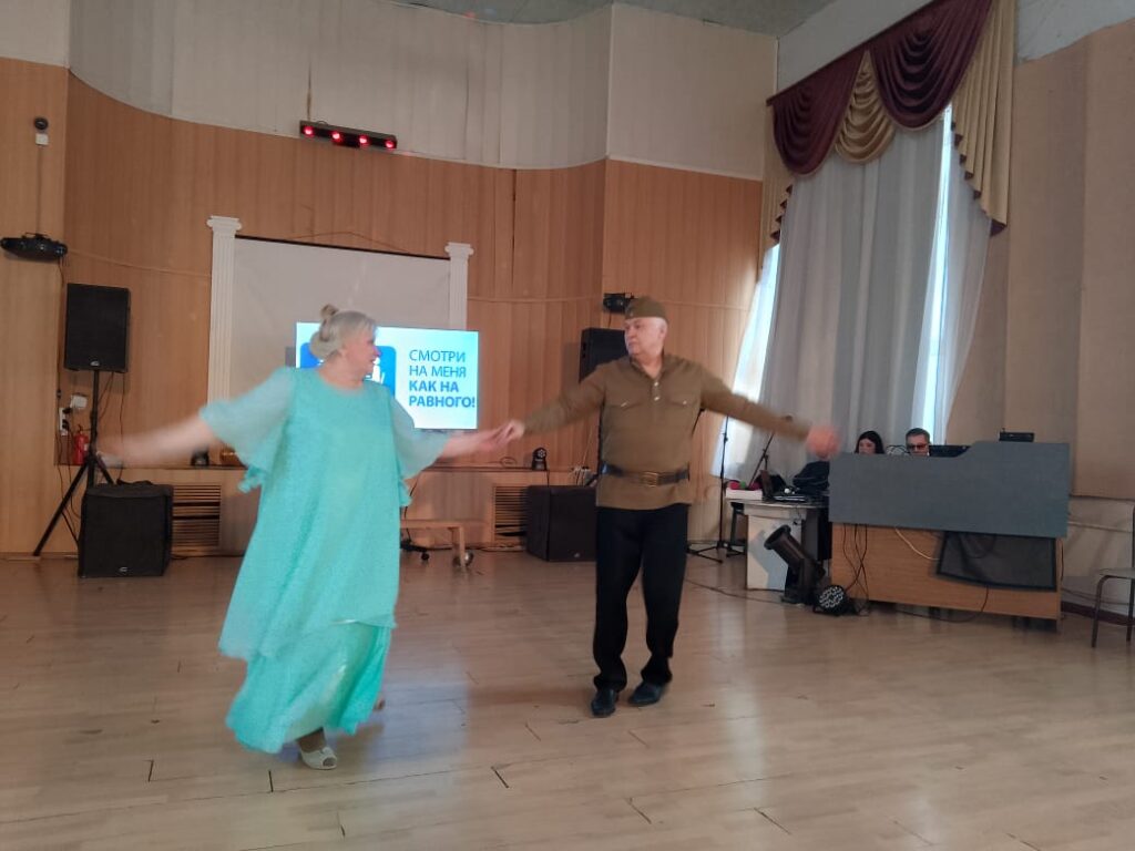 Танцевальная пара выступает в зале: мужчина в военной форме и женщина в бирюзовом платье. Их движения передают настроение послевоенного времени — облегчение и надежду на мирную жизнь.