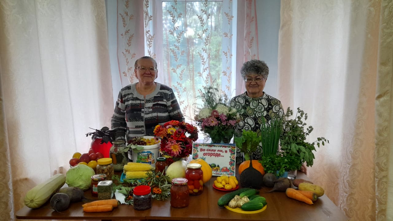 две участницы позируют за столом с богатым урожаем: кабачки, морковь, свёкла, кукуруза, зелень, банки с домашними заготовками, а также букеты цветов.