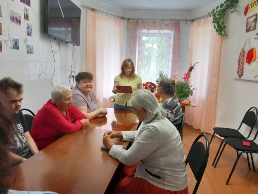 момент мероприятия: участники сидят за столом, ведущая с листом в руках читает текст, а рядом стоит праздничная выставка овощей и цветов.