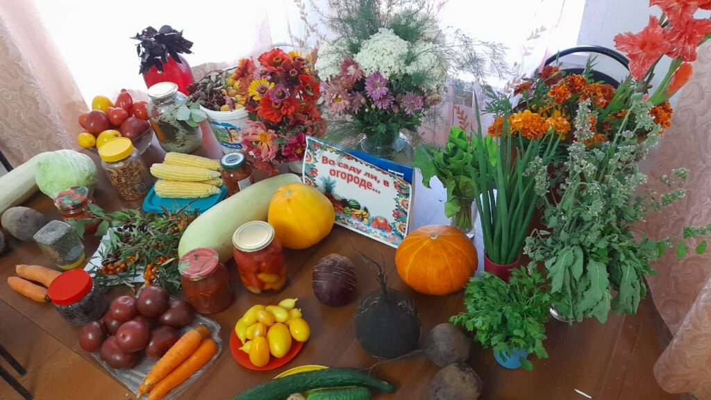 крупный план выставочного стола: тыквы, морковь, огурцы, помидоры, кукуруза, банки с компотами и соленьями, букеты бархатцев и гладиолусов, зелень и травы.