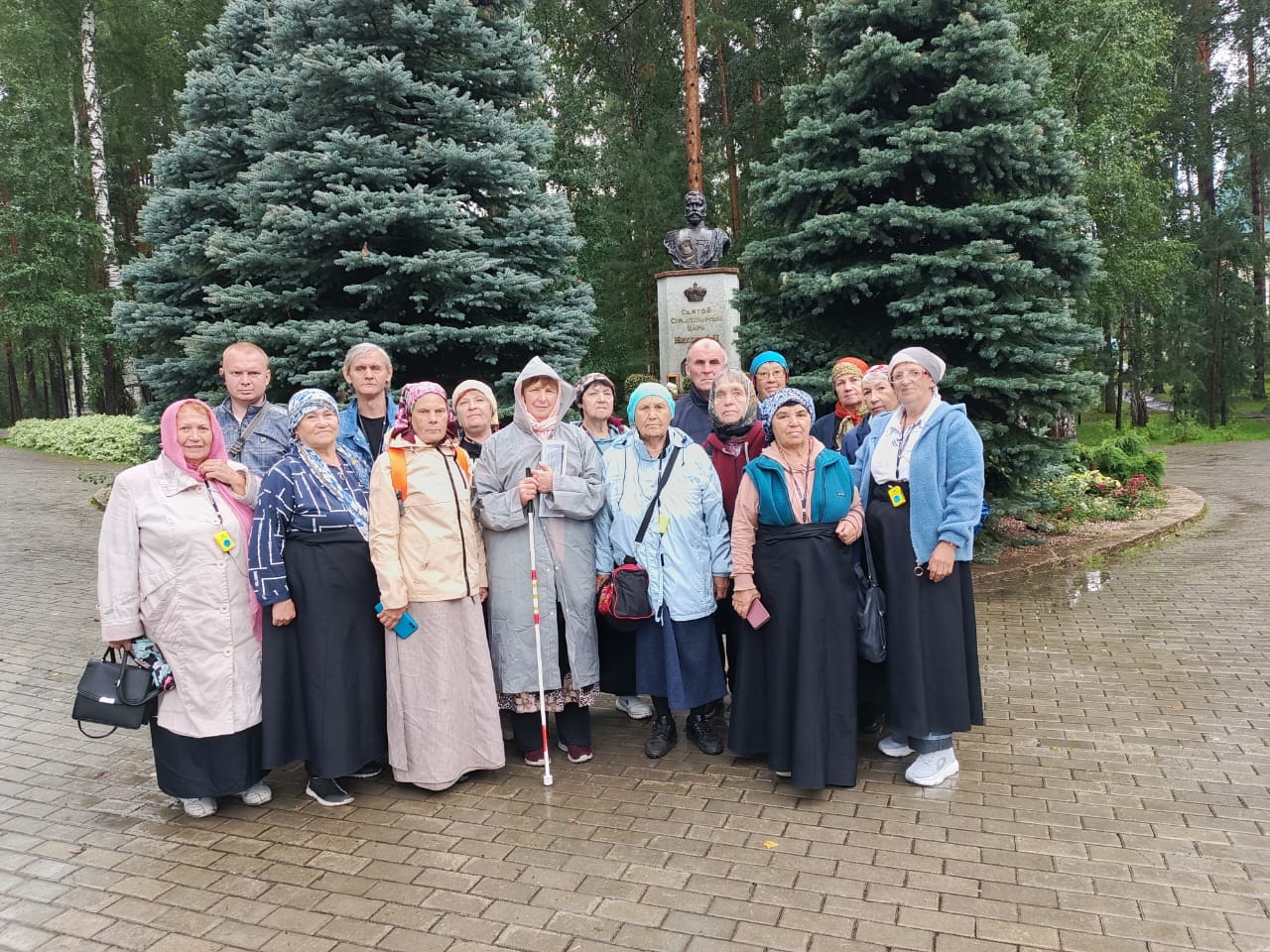 На фотографии — группа участников досугового центра «Огонёк» Верхнеуфалейской МО ВОС во время экскурсии в Ганину Яму. Люди стоят вместе у памятника на территории монастыря, на фоне зелёных елей. Участники одеты по паломнической традиции: женщины в юбках и платках, некоторые с тростями для слабовидящих, на груди у многих видно оборудование для тифлокомментирования.