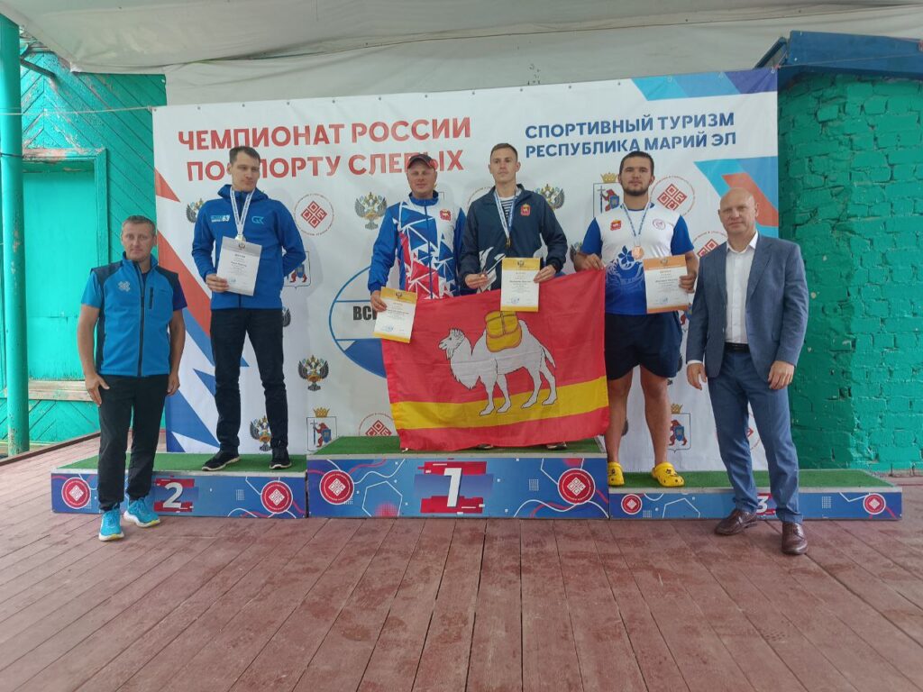 На пьедестале победителей стоят мужчины-спортсмены с наградами и дипломами. В центре — спортсмены Челябинской области с флагом региона.
