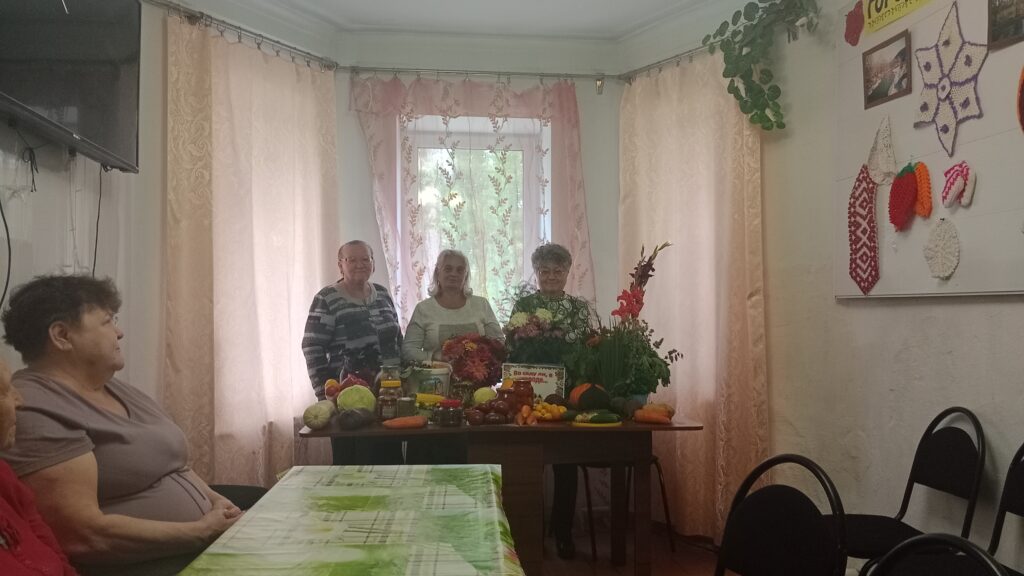 несколько женщин стоят за праздничным столом, украшенным овощами, фруктами, цветами и заготовками. На переднем плане – вывеска «Во саду ли, в огороде…».