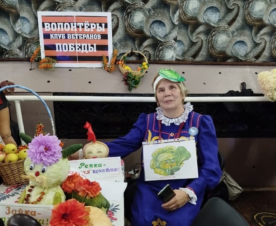 Валентина Митрохина у выставочного стола команды «Волонтёры Победы» с композициями «Редька-милая кокетка» и «Зайка-зазнайка».