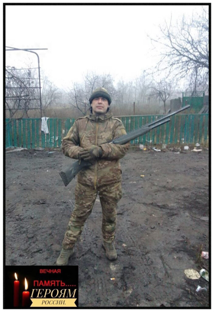 Мужчина в камуфляжной военной форме стоит на грязной земле на открытом участке, фон – редкие деревья без листьев и низкий забор. Он держит в руках большое ружьё и смотрит в камеру. На нём шапка и перчатки. В левом нижнем углу размещён траурный уголок с чёрным фоном, изображением двух зажжённых свечей и надписью:

ВЕЧНАЯ
ПАМЯТЬ…
ГЕРОЯМ
РОССИИ.