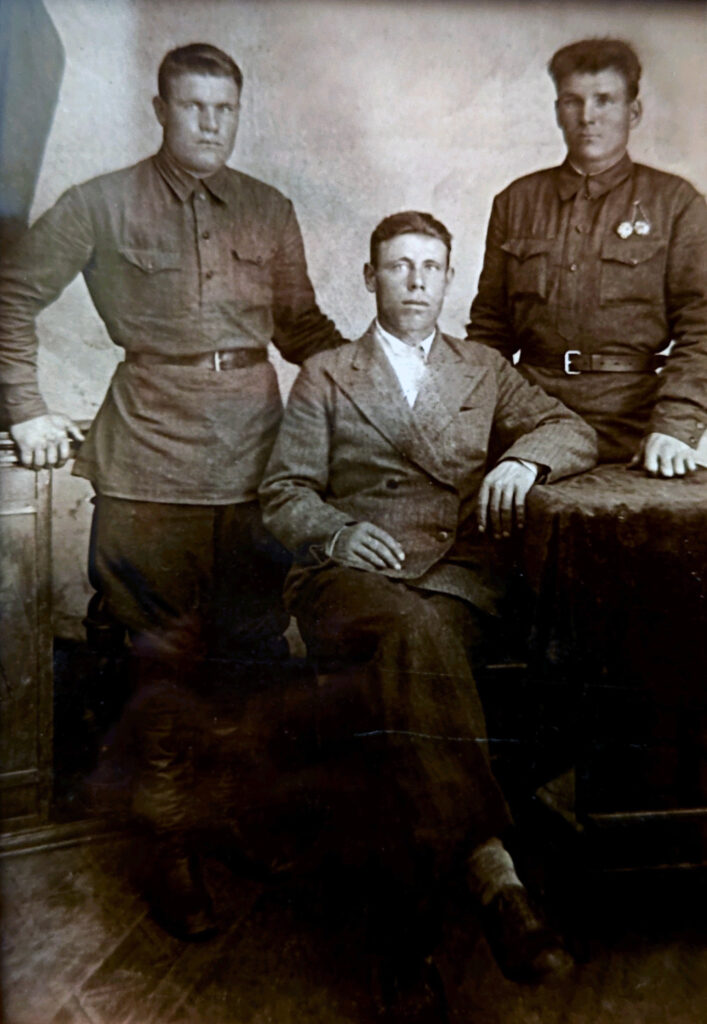 Перед отправкой на фронт, фотография 1942 года.
На cтарой черно-белой фотографии трое мужчин. Двое из них стоят по бокам — они одеты в военную форму с ремнями, на груди одного из них видны медали или знаки отличия. Третий мужчина сидит между ними на стуле и одет в светлый двубортный костюм. У всех серьезные, спокойные выражения лиц. На заднем плане нейтральный фон, виден стол с темной скатертью, на который опирается один из мужчин. 
