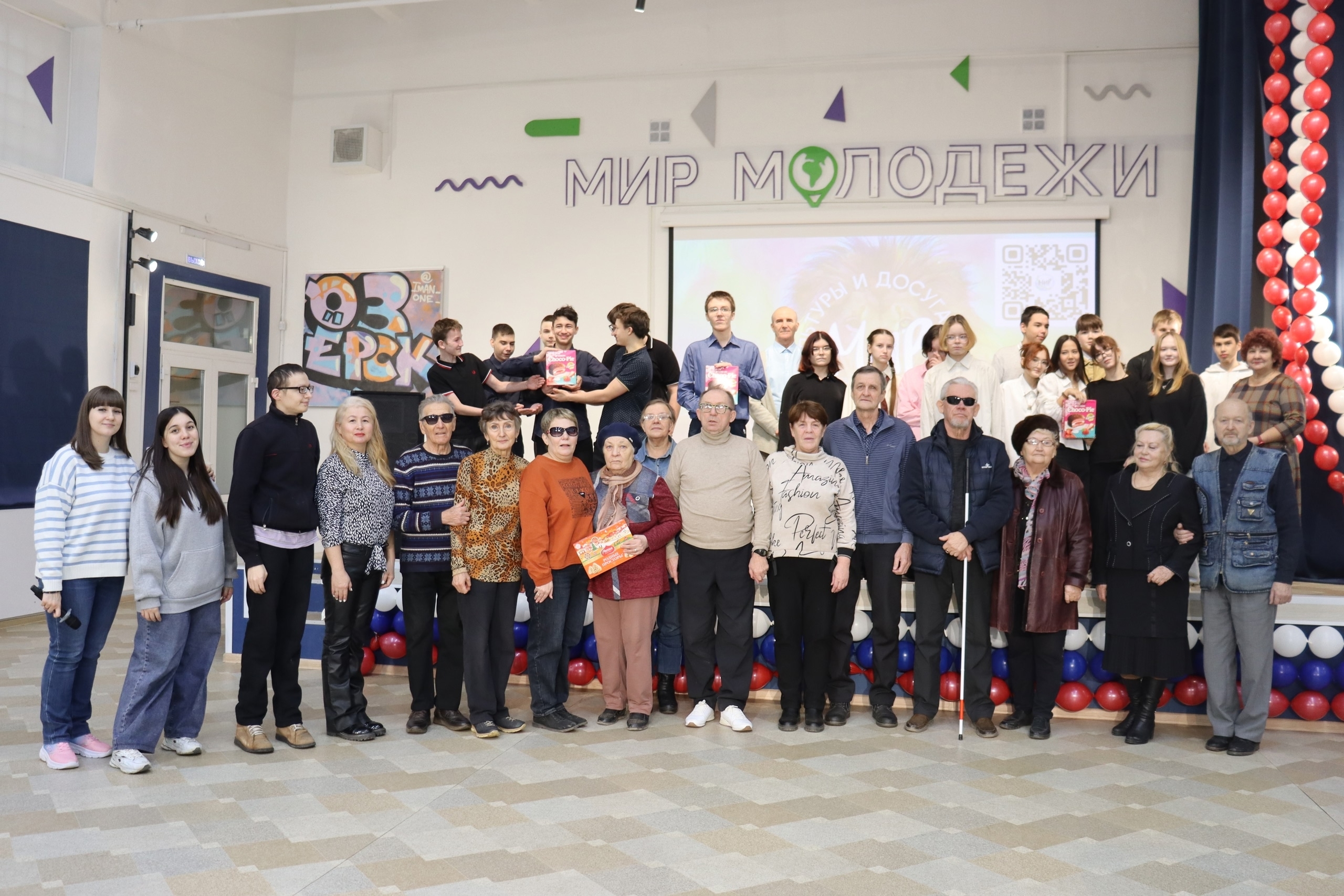 Групповое фото в помещении На фото большая группа людей разного возраста стоит и сидит, позируя для совместного снимка. Люди расположены в несколько рядов — впереди взрослые и пожилые, сзади молодежь и подростки. Некоторые держат в руках коробки или подарки. Помещение украшено воздушными шарами красного, белого и синего цветов — они составляют гирлянды вдоль сцены. На задней стене висит надпись "МИР МОЛОДЕЖИ", а под ней — экран с презентацией и QR-кодом, а также надпись на экране "КУЛЬТУРЫ И ДОСУГА". Справа на заднем плане часть людей стоит на ступенях сценической площадки. Атмосфера торжественная и дружеская, у большинства людей спокойные или радостные лица.