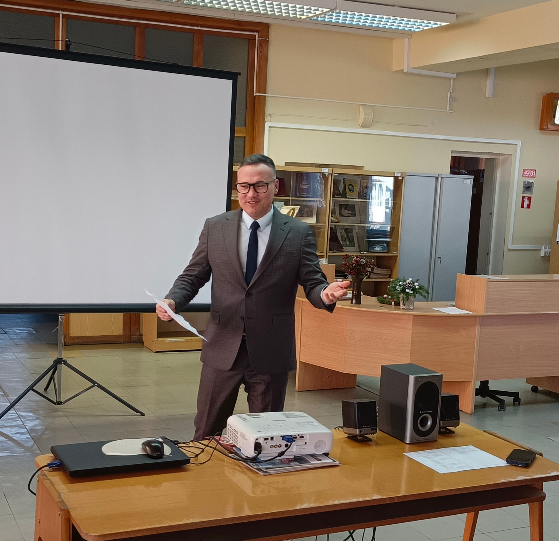 Мужчина в деловом костюме стоит перед большим белым экраном для проектора в помещении библиотеки. Он держит бумагу в левой руке и улыбается. На переднем плане — деревянный стол, на котором стоят ноутбук, проектор, колонки и мышь. В помещении мебель светлого дерева, на заднем плане стеллажи с папками, цветы, видны коридоры и двери. Атмосфера формальная и деловая.
