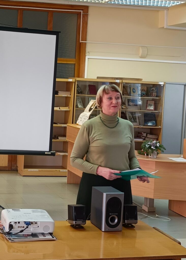Женщина выступает перед собравшимися. На столе перед ней проектор и колонки, за спиной стеллажи с книгами.