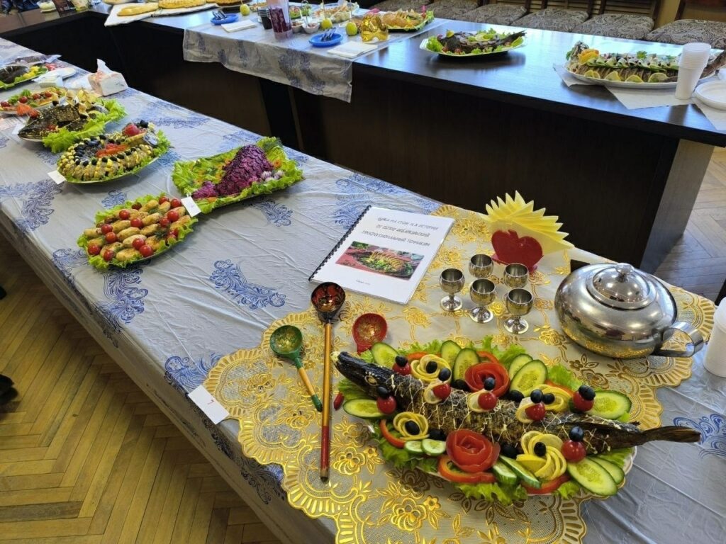 Праздничный стол с блюдами. На переднем плане — большое блюдо с фаршированной рыбой, украшенной кружочками лимона, огурца, помидорами черри и маслинами, а также фигурками из овощей в виде роз. Рядом стоит металлический чайник и шесть металлических рюмочек. Перед чайником лежат две расписные ложки. На салфетке лежит раскрытая брошюра с текстом и фотографией блюда и декоративное украшение из нарезанных ломтиков яблока, выполненное в форме веера.
Дальше по столу стоят ещё блюда с закусками, часть из которых украшена овощами, зеленью и ягодами