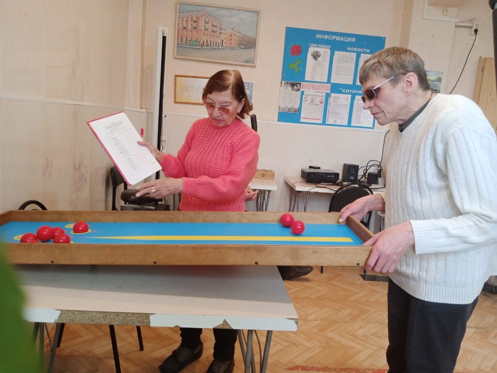 Женщина в розовом свитере держит в руке папку с бумагами и указывает на что-то внутри неё. Рядом с ней стоит мужчина в белом свитере и тёмных очках, он держит деревянную рамку с голубым игровым полем и красными шарами — это настольная игра для людей с нарушением зрения. На стене за ними висит синяя информационная доска, на столе стоит техника и другие предметы. На стене также висят картины. Пол — паркетный.