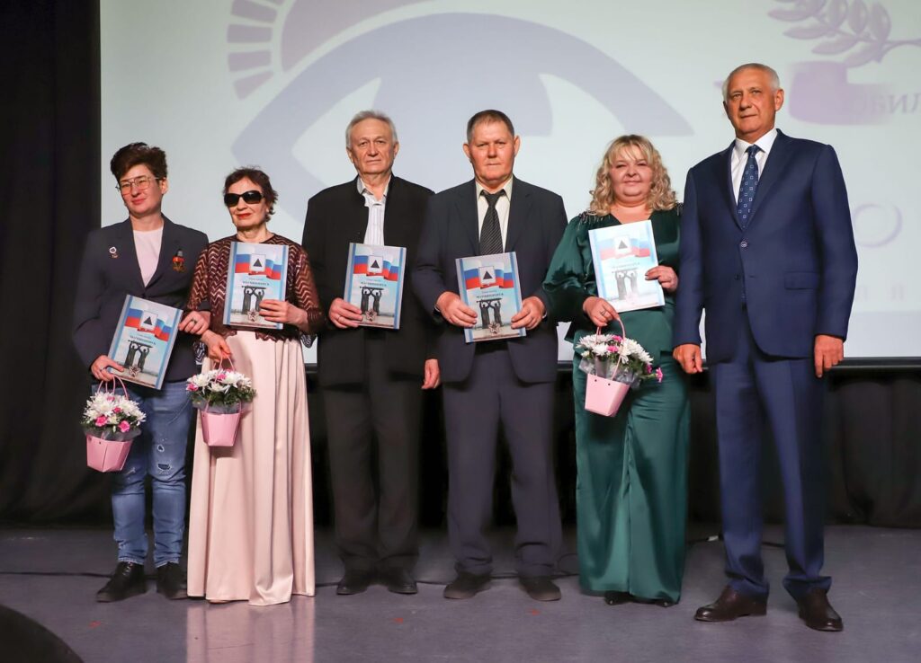 Шесть человек на сцене, часть из них держит папки и цветы. Официальное мероприятие, люди нарядно одеты, на заднем плане логотип на экране.