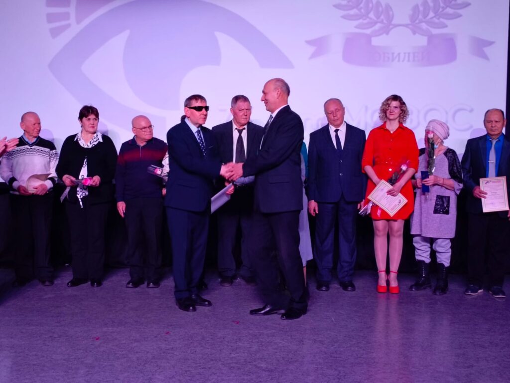 Группа людей на сцене, двое мужчин в центре жмут друг другу руки. Остальные держат грамоты и цветы. Официальная церемония награждения, фон с изображением глаза и надписью "Юбилей".