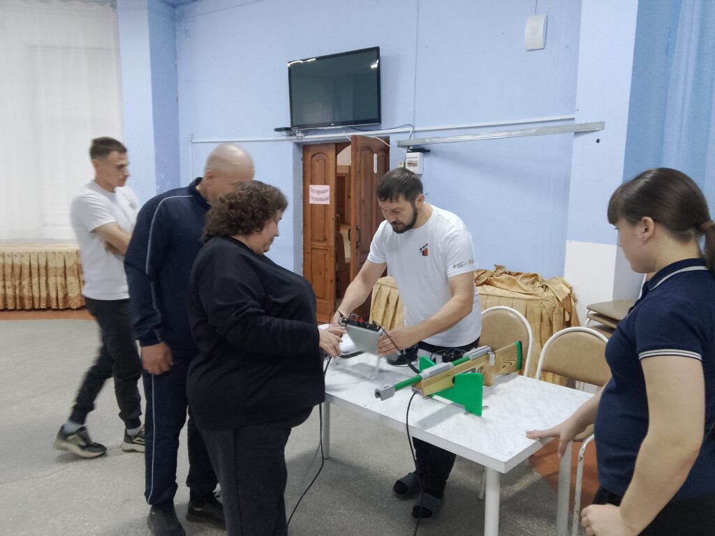 Группа людей у стола внутри помещения, один человек обучает другого пользоваться винтовкой на подставке, остальные наблюдают.