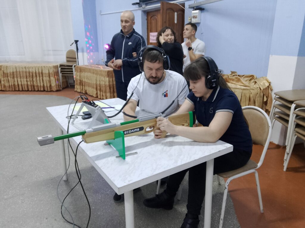 Женщина и мужчина за столом осваивают электронный тир, оба в наушниках, мужчина помогает прицелиться. На заднем плане несколько наблюдателей.