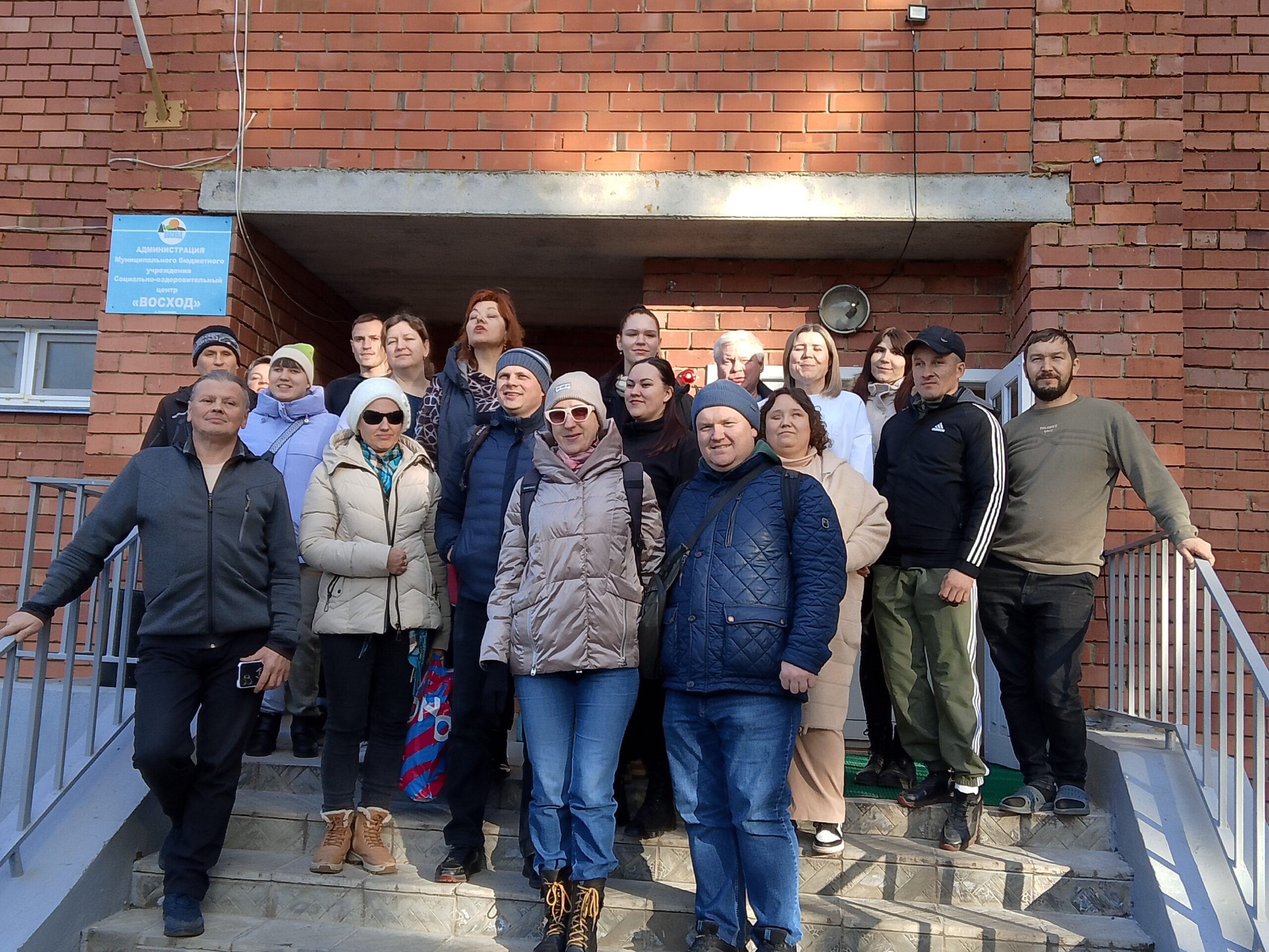 Группа из 18 человек стоит на ступенях у входа в кирпичное здание, одеты по-осеннему, на стене табличка "Восход".