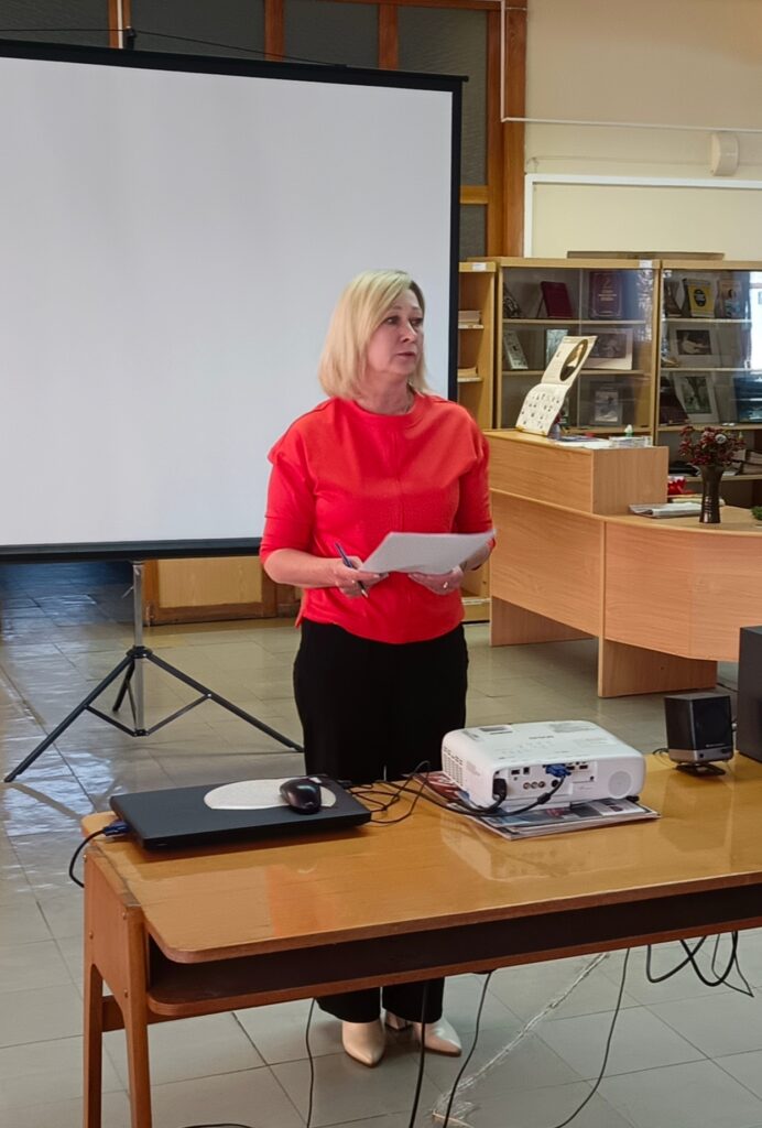 Женщина в красной кофте стоит у экрана, держит бумаги, перед ней ноутбук и проектор. 