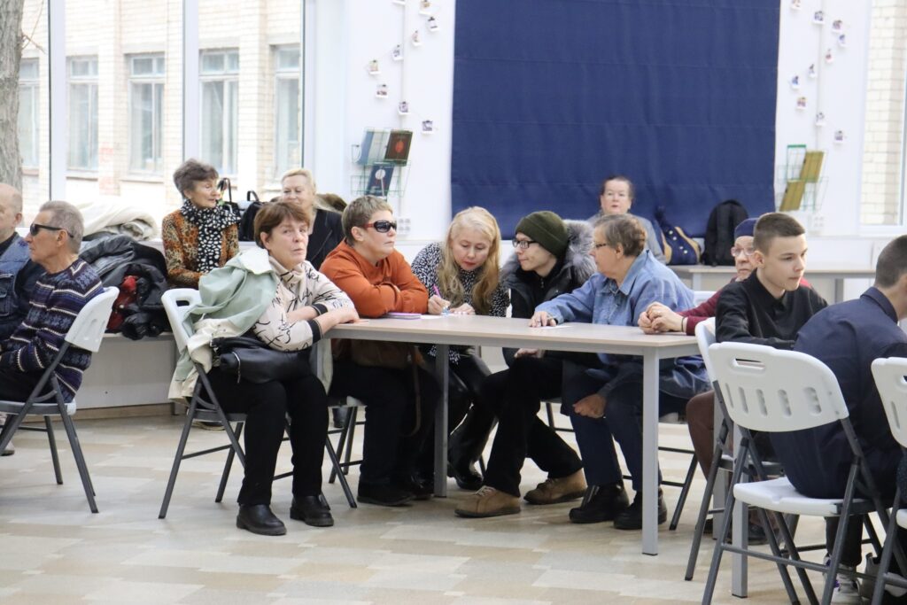 Группа людей, сидящих за столами в ярко освещённой комнате. На переднем плане за длинным прямоугольным столом расположились люди разного возраста — женщины и мужчины. Одна женщина пишет что-то в тетради, остальные наблюдают или обсуждают. В зале присутствуют как молодёжь, так и пожилые люди. Слева и сзади видны большие окна, пустые кресла и настенный декор.
