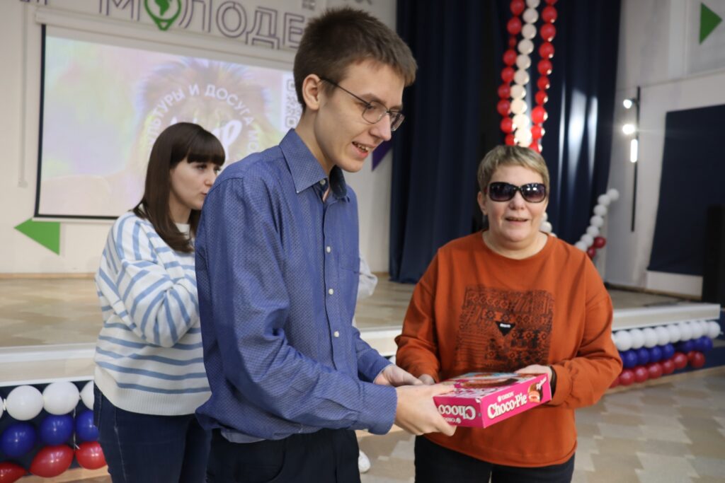 Три человека в помещении. На переднем плане молодой человек в синей рубашке держит коробку с надписью "Choco Pie", рядом с ним женщина в оранжевом свитшоте и темных очках- председатель озёрской мо вос татьяна решетникова, она также держит коробку за край. На заднем плане стоит девушка в полосатом свитере. Оформление зала праздничное — гирлянды из красных, белых и синих шаров, на сцене висит большой экран с надписями на русском языке. 