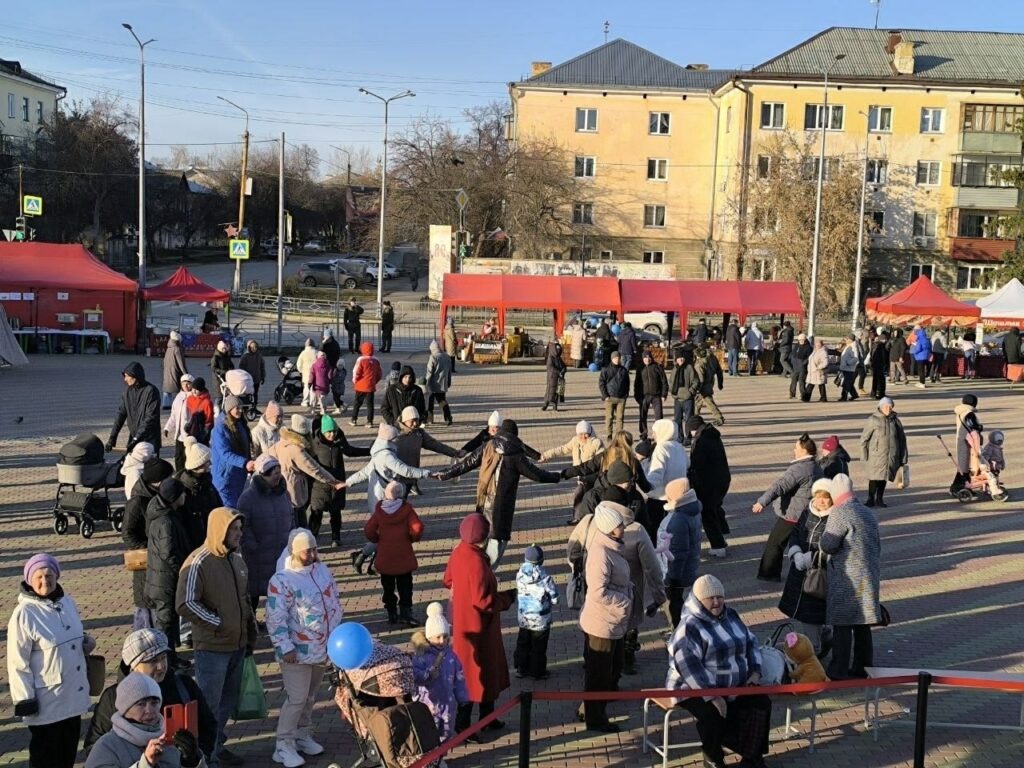 Площадь с большим количеством людей в зимней одежде. На переднем плане несколько человек с детскими колясками, кто-то держит синий воздушный шарик. В центре снимка – люди стоят в кругу, держась за руки, возможно, танцуют хоровод. Позади них – торговые ряды с красными тентами, где идет ярмарка, можно увидеть прилавки с товарами и продавцов. На заднем плане – жилые дома и припаркованные машины, а также деревья без листвы. Мероприятие проходит на открытом воздухе при солнечной погоде.