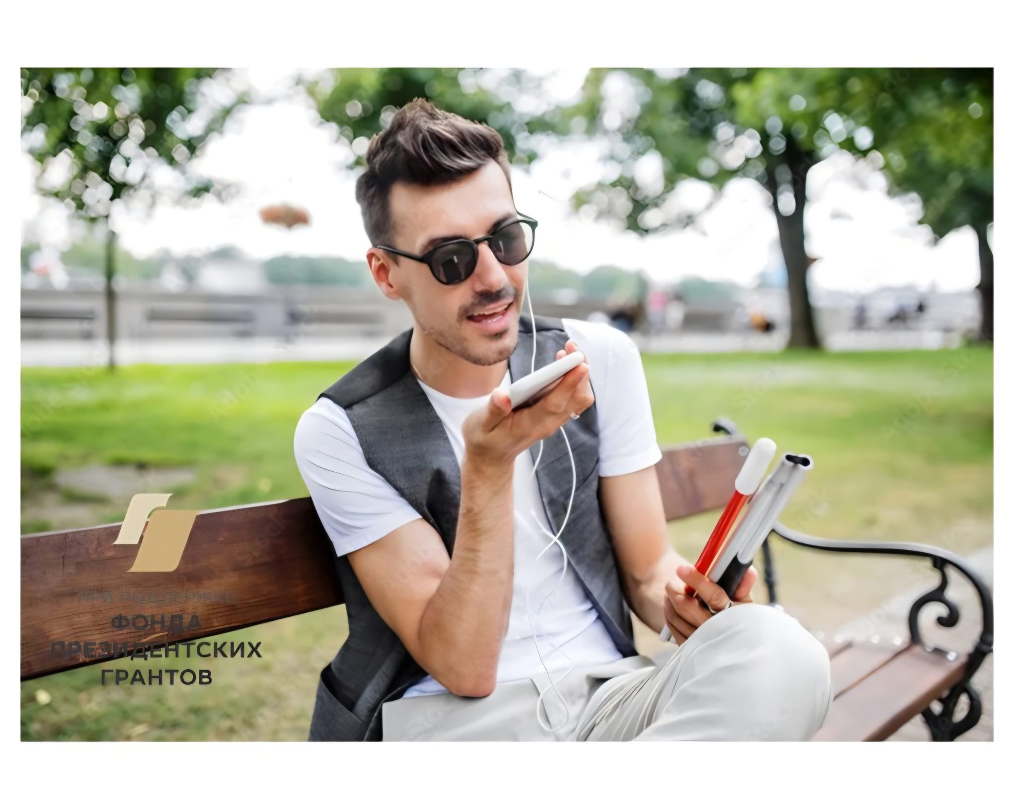 Молодой мужчина с короткими тёмными волосами и усами сидит на деревянной скамейке в городском парке. На нём солнечные очки, белая футболка и серый жилет. Он держит в одной руке складную белую трость для незрячих, а в другой — смартфон, на который, вероятно, что-то диктует или слушает голосовое сообщение. Из смартфона идут наушники. Фон — зелёная трава, деревья и немного размытые объекты на заднем плане. В левом нижнем углу логотип с надписью: "ФОНД ПРЕЗИДЕНТСКИХ ГРАНТОВ".