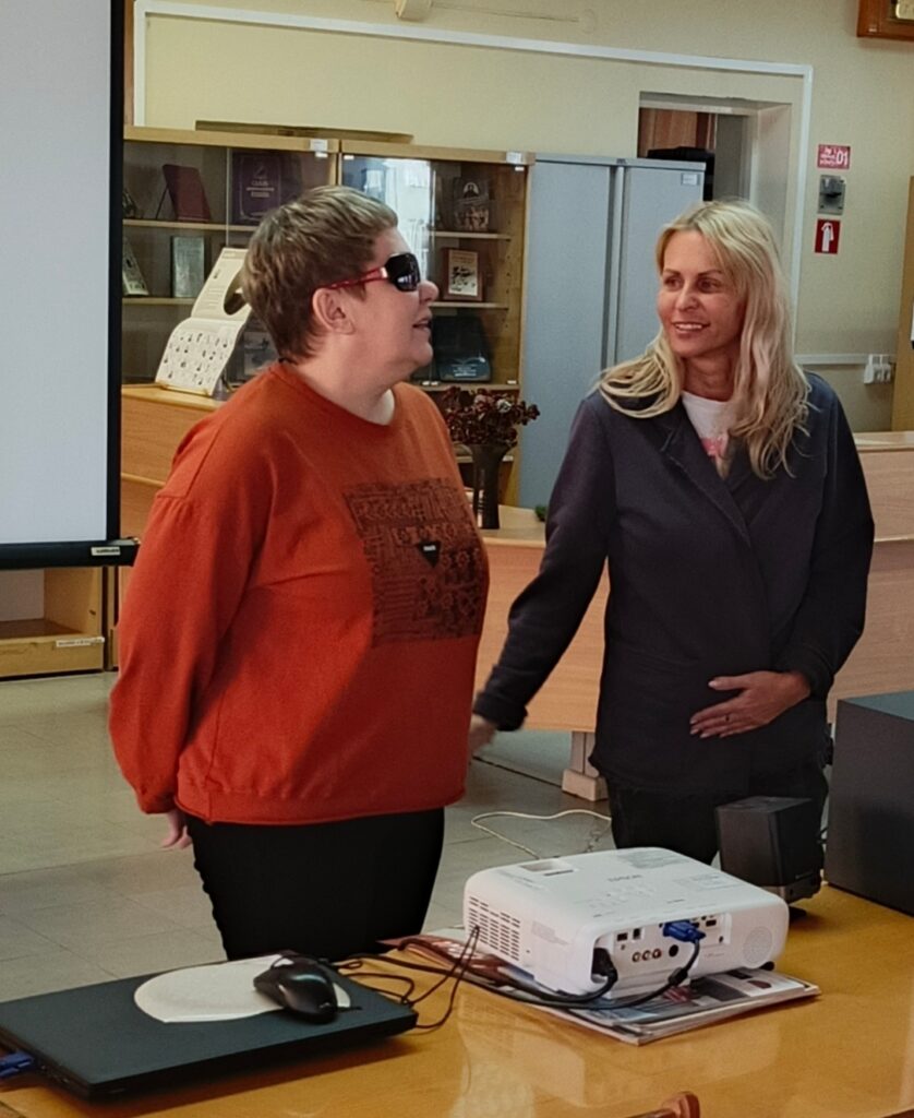 Две женщины стоят рядом за столом с проектором и ноутбуком в помещении библиотеки. Одна женщина в тёмных очках и оранжевом свитере, другая — светловолосая и в тёмной куртке.