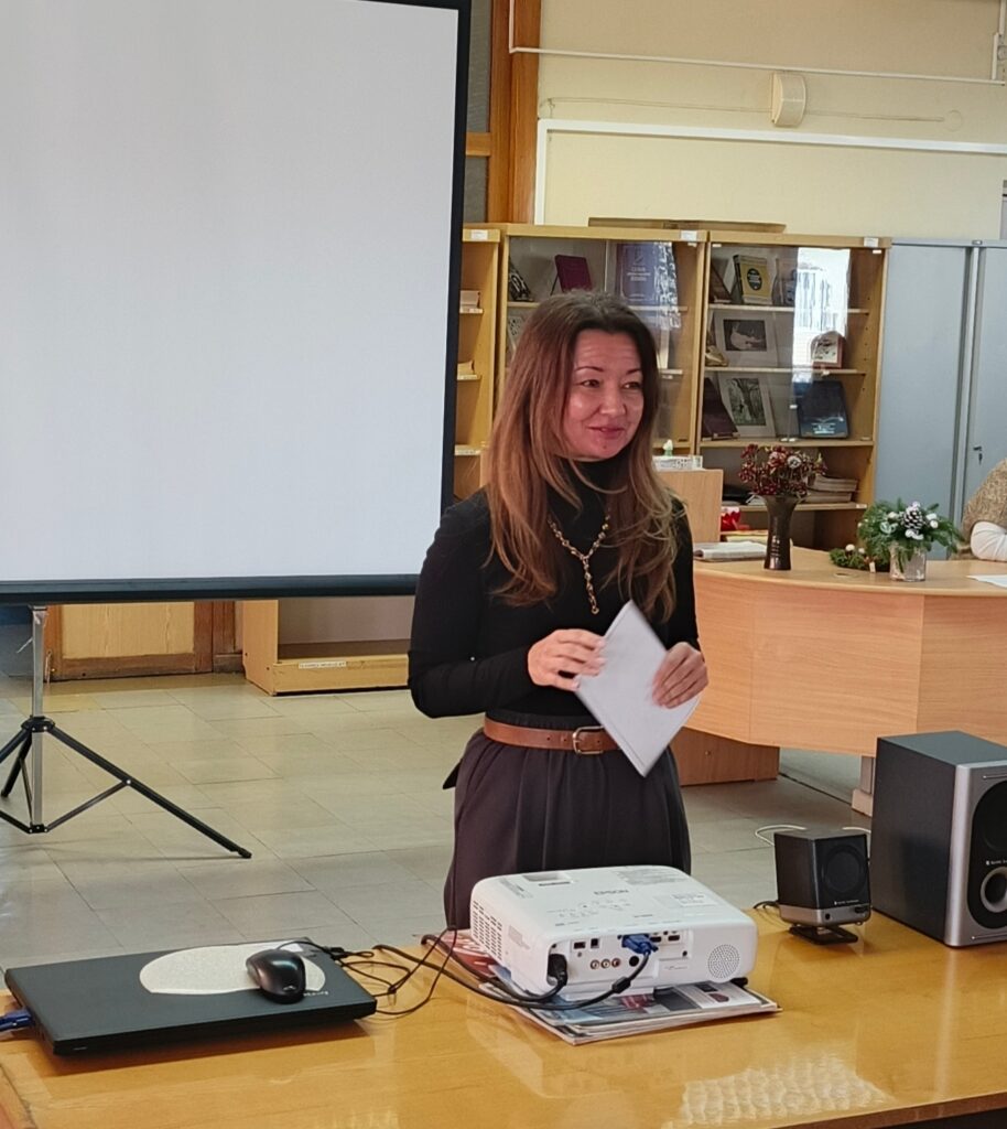 Женщина стоит у стола с проектором и ноутбуком, держит лист бумаги, за ней экран для презентации и книжные стеллажи.