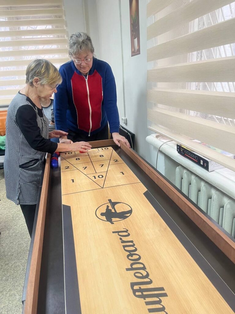 Две женщины играют в шаффлборд, одна передвигает фишку, другая наблюдает. Стол с разметкой очков, рядом лежит игра "Катан". Светлая комната с жалюзи.