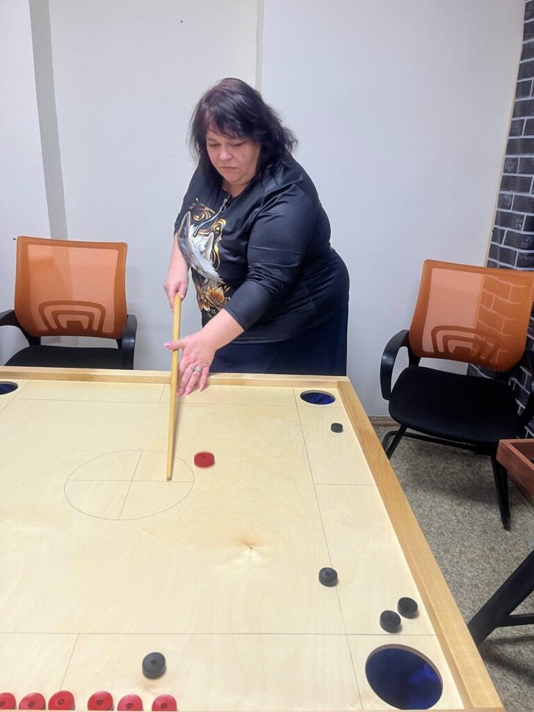 Женщина играет в настольную игру за столом, держит кий, на столе фишки, вокруг стоят офисные кресла.