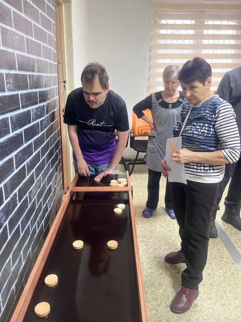 участники играют в настольную игру с деревянными фишками. Мужчина бросает фишку, две женщины наблюдают.