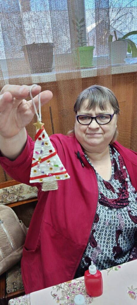 Женщина держит в руке новогоднюю поделку-ёлочку из ткани, сидит за столом,