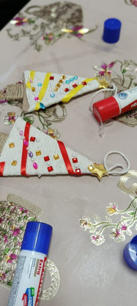 Две самодельные ёлочки из картона, обмотанные ниткой, украшены стразами и ленточками. Рядом клей и скатерть с вышивкой.