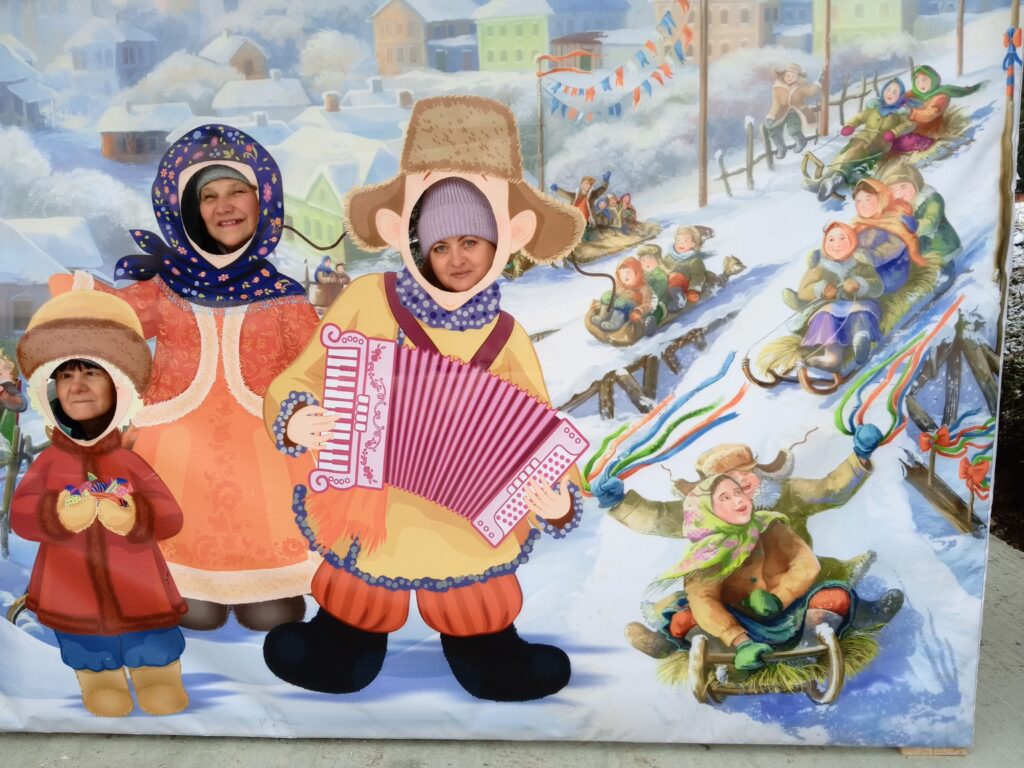 Три женщины вставили лица в вырезы яркой зимней фотозоны, изображающей традиционных персонажей на фоне зимней горки с катающимися на санках людьми.