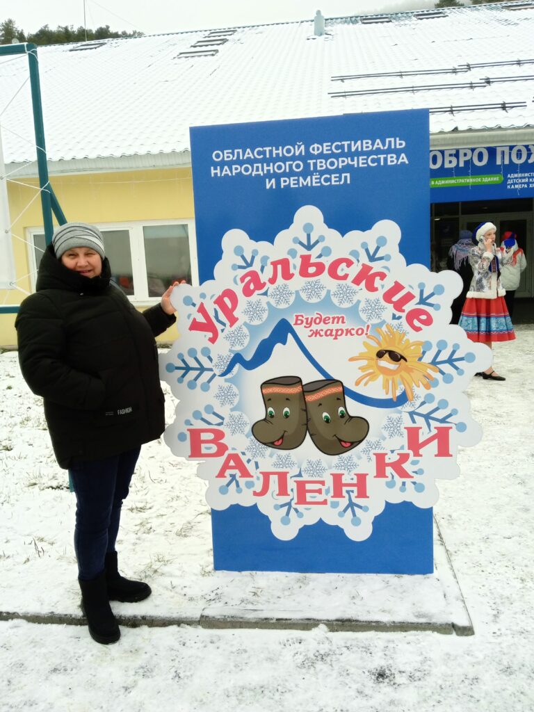 Женщина стоит у щита с надписью "Уральские валенки" на фестивале народного творчества, вокруг зима и снег.