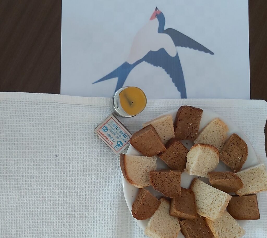 Тарелка с кусочками белого и чёрного хлеба на белой салфетке; рядом маленькая жёлтая свеча в стекле и коробок спичек; на фоне рисунок летящей птицы.