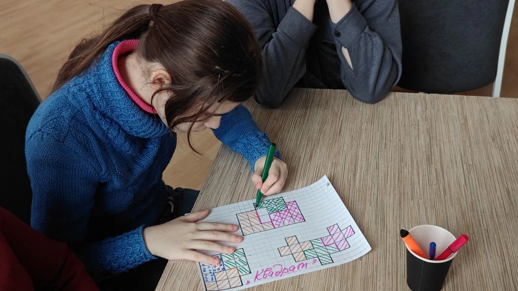 Девушка за столом раскрашивает зелёным маркером клетчатый лист с геометрическими фигурами; рядом стаканчик с фломастерами. На листе внизу написано: квадрат