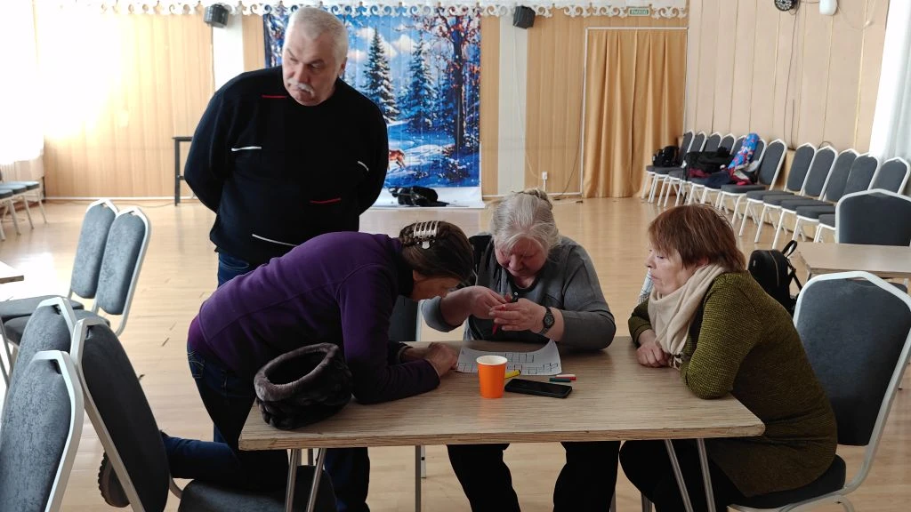 В зале за столом трое женщин вместе рассматривают задание на листе, рядом стоит мужчина и наблюдает; вокруг ряды стульев, на столе стакан и телефон.