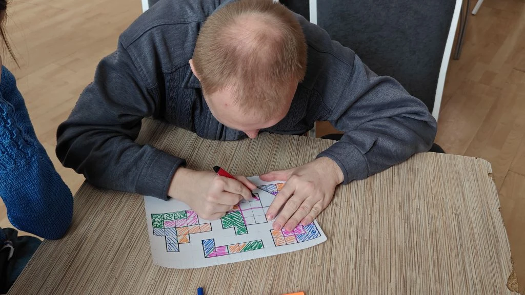 Мужчина за столом раскрашивает фломастером лист с геометрическим рисунком/головоломкой из клеток и фигур