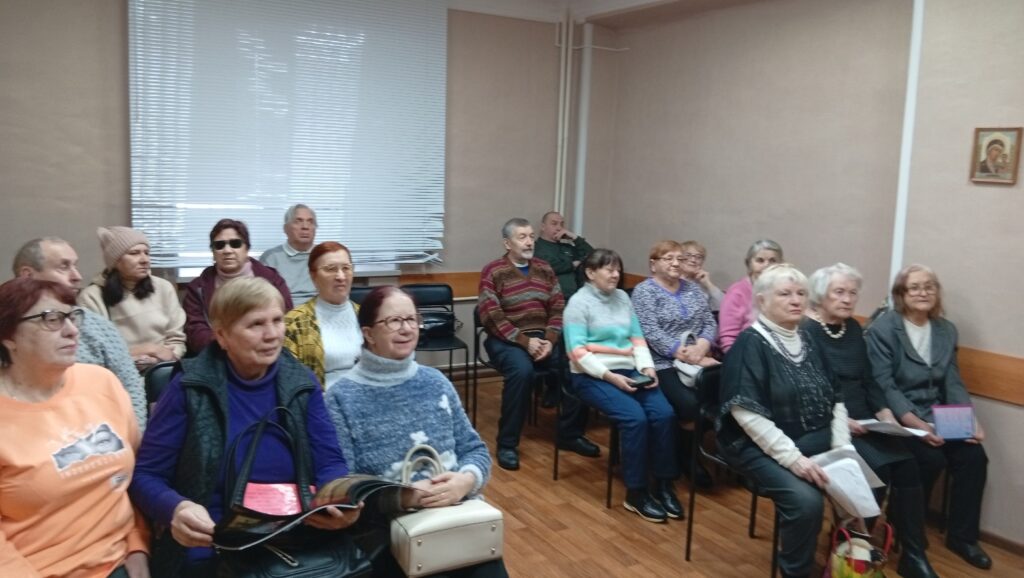 Зрители, в основном пожилые, сидят рядами в небольшой комнате для собраний/ожидания; слева окно с жалюзи, у некоторых в руках документы и сумки.